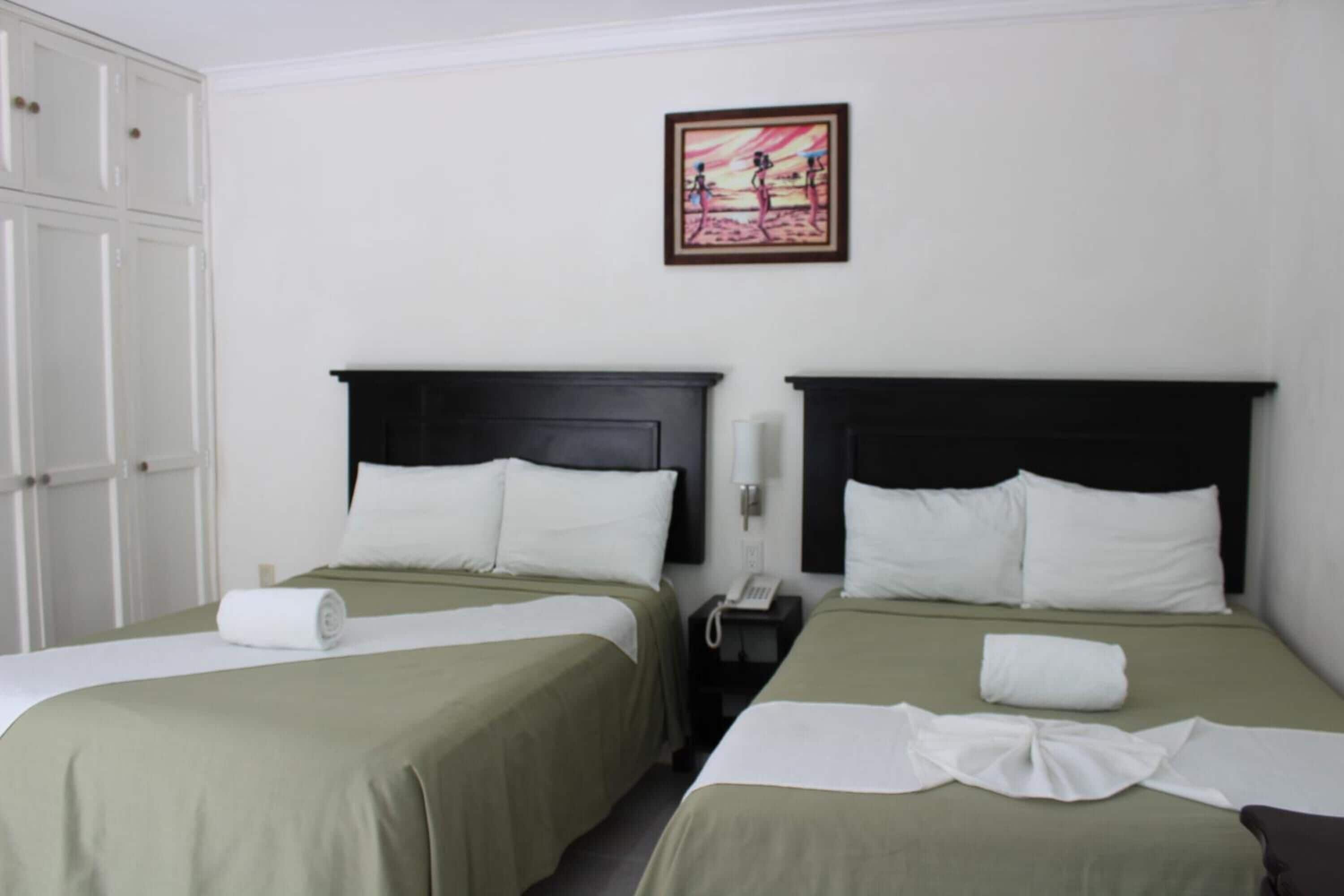 Une chambre d’hôtel avec deux lits, chacun doté d’une tête de lit sombre et de draps blancs. Un tableau est accroché au mur au-dessus des lits, un téléphone se trouve sur la table de chevet et une porte blanche est située à gauche.