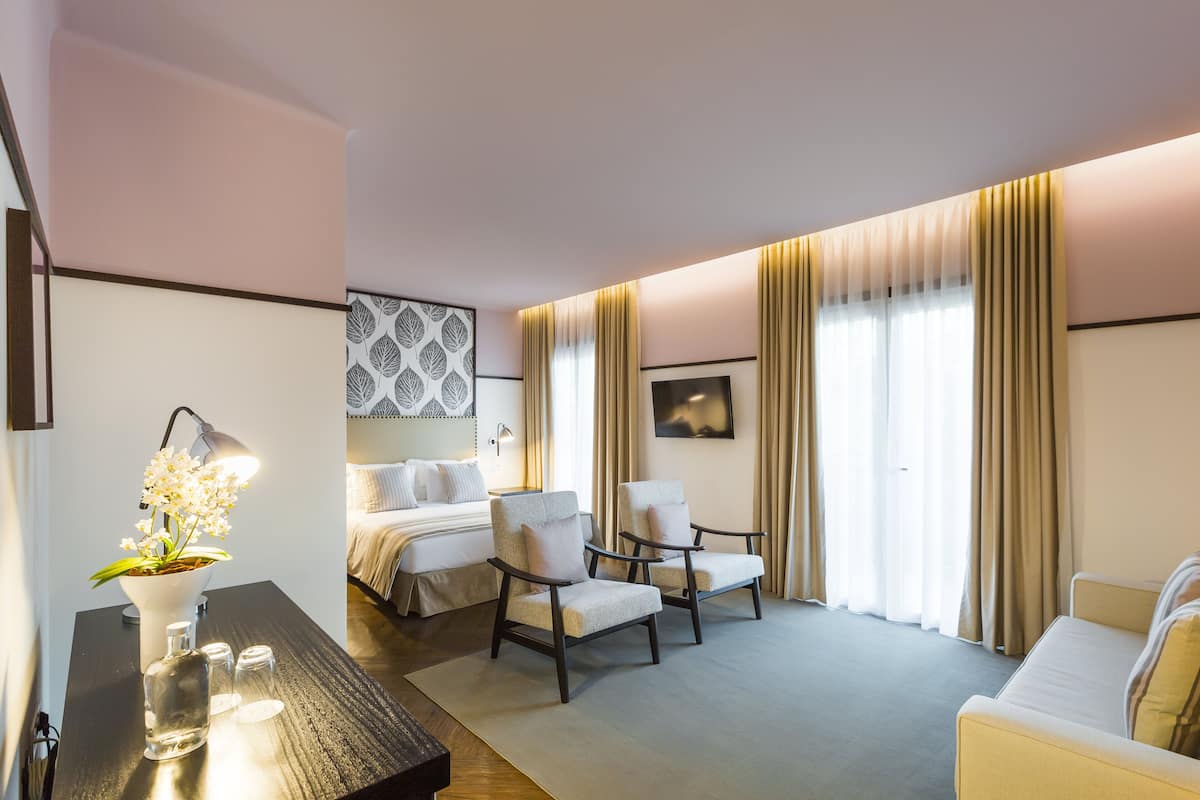 A hotel room with a bed, two armchairs, a desk with a lamp, and a vase with flowers.