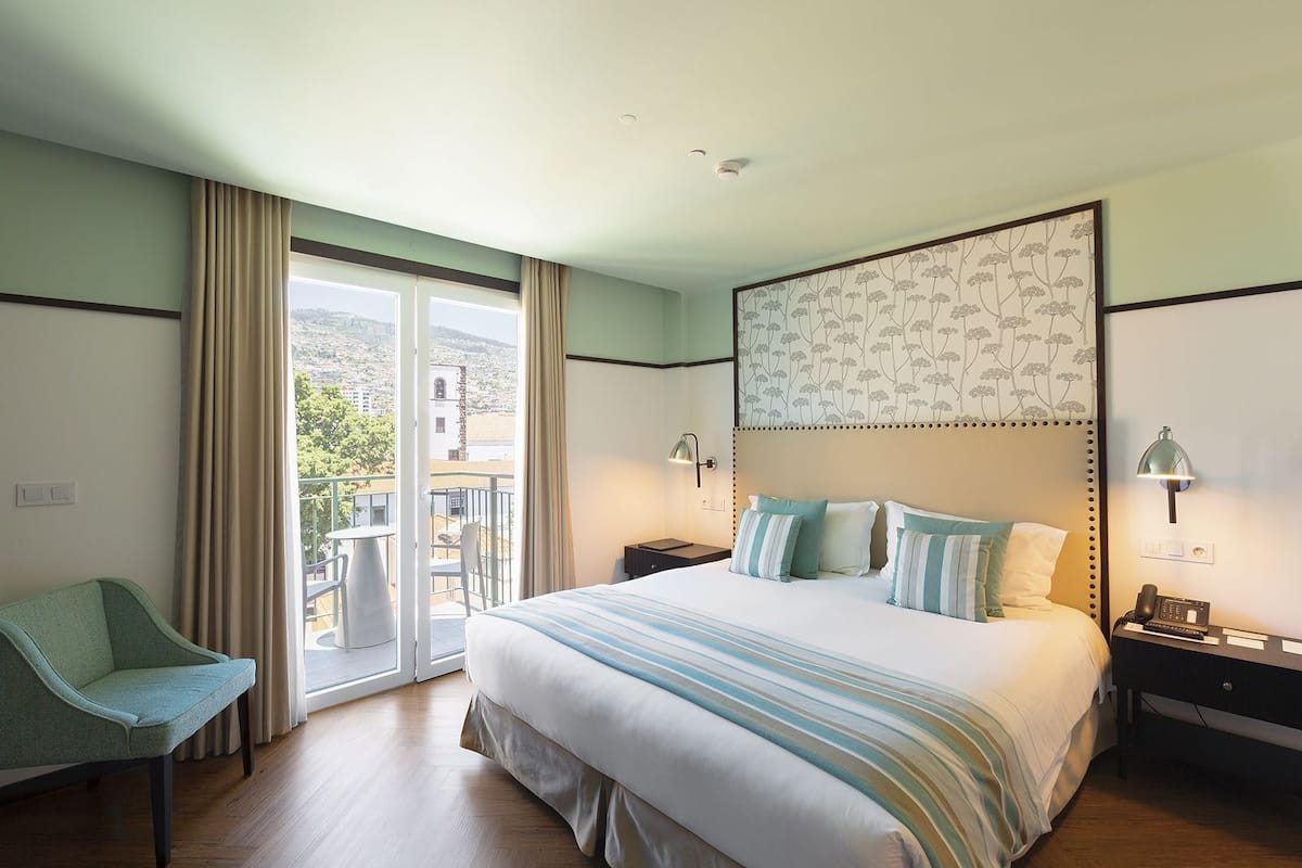 A hotel room with a bed, a chair, a nightstand, a phone, and a view of the outside.