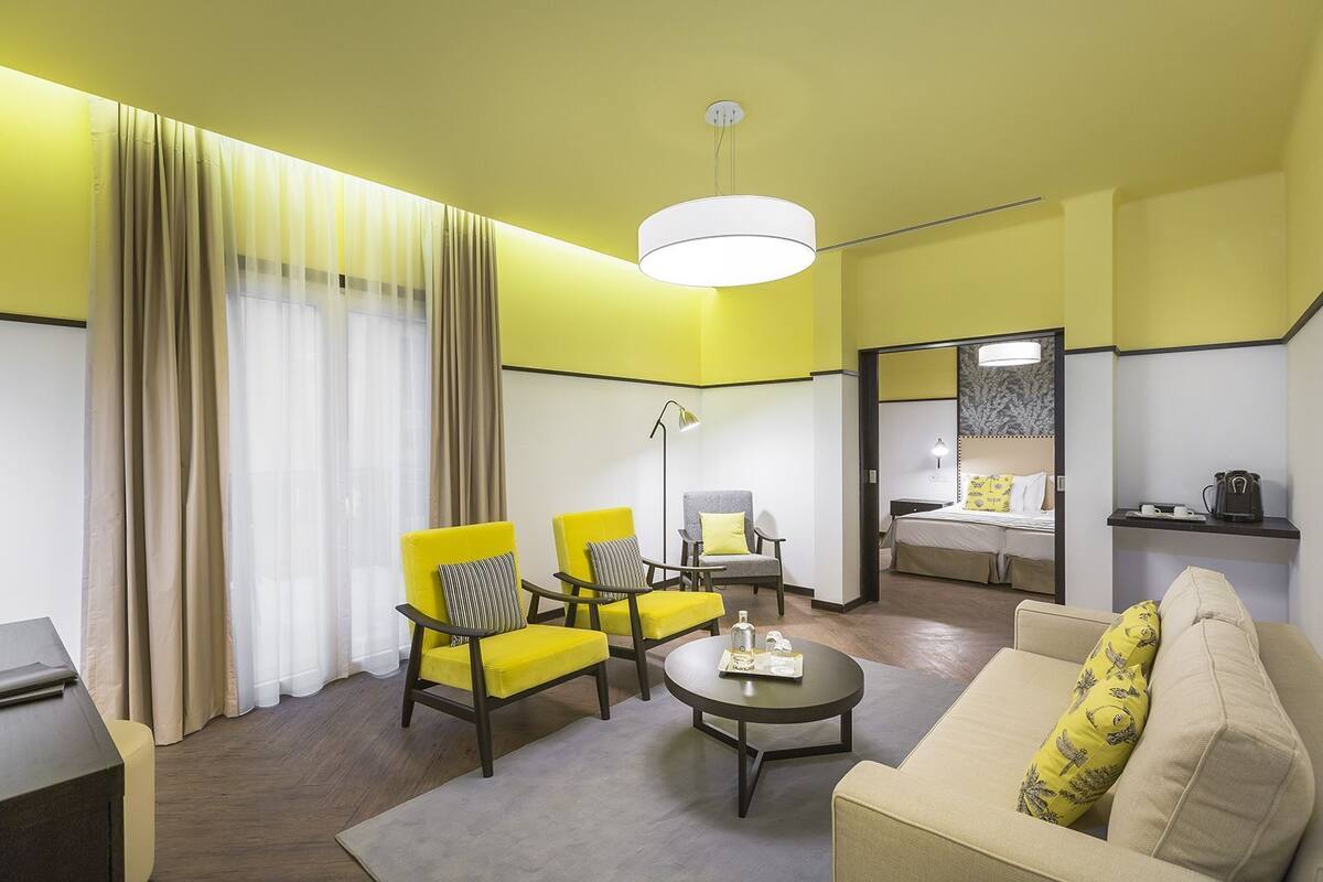 A modern hotel room with a yellow and white color scheme, featuring a sofa, two armchairs, a coffee table, a desk, and a bed with a nightstand.