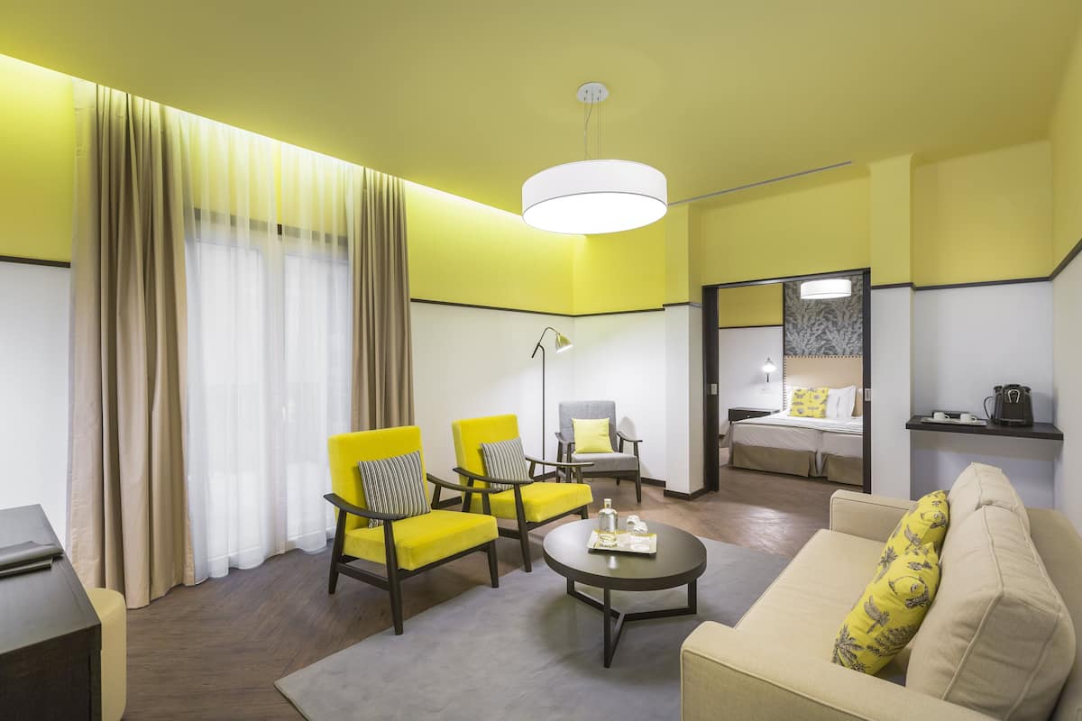 A hotel room with yellow walls, a sofa, two yellow armchairs, a coffee table, a bed, a mirror, and a desk.