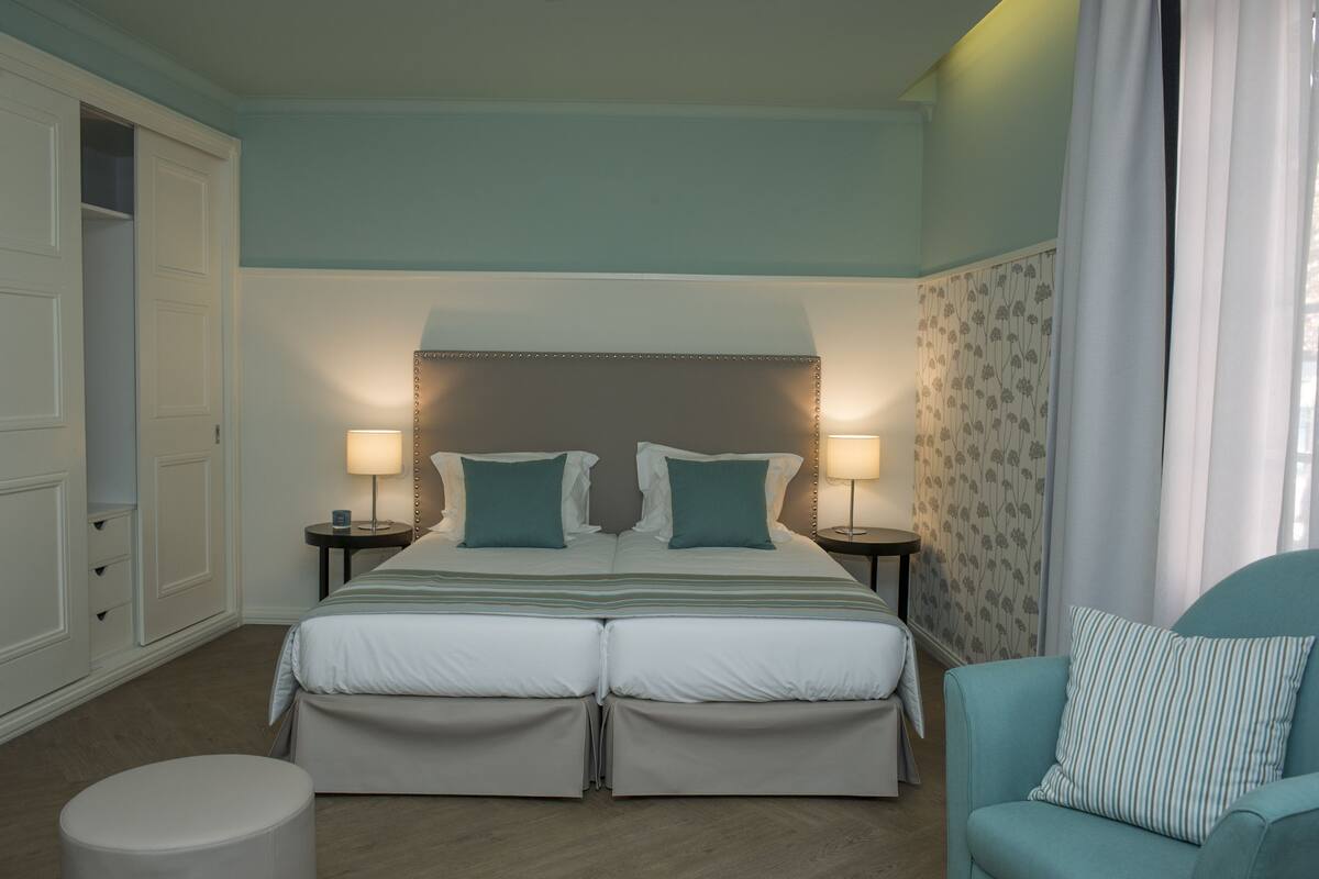 A double bed with two bedside tables, a chair, a round ottoman, and a wardrobe.
