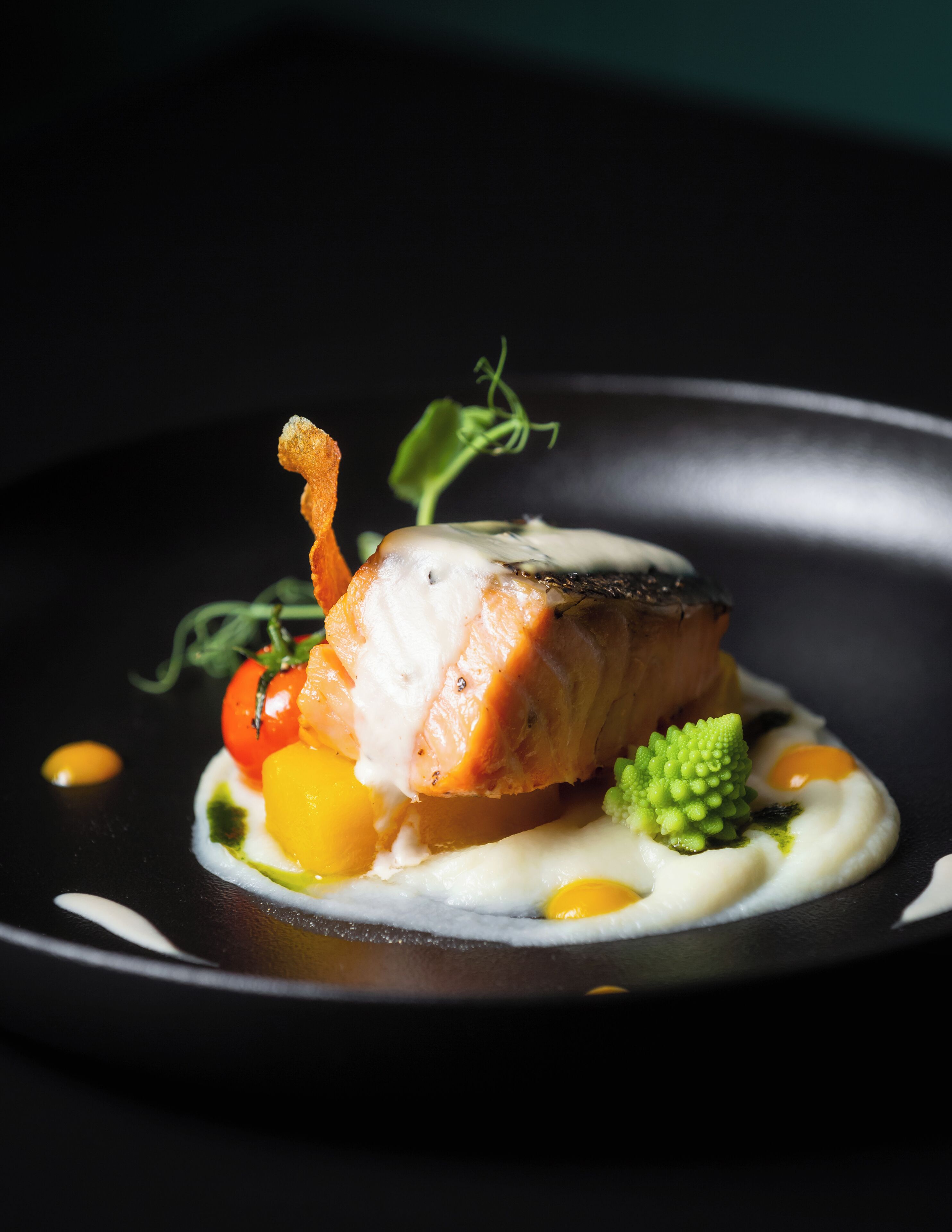 A dish with salmon, vegetables, and sauce on a black plate.
