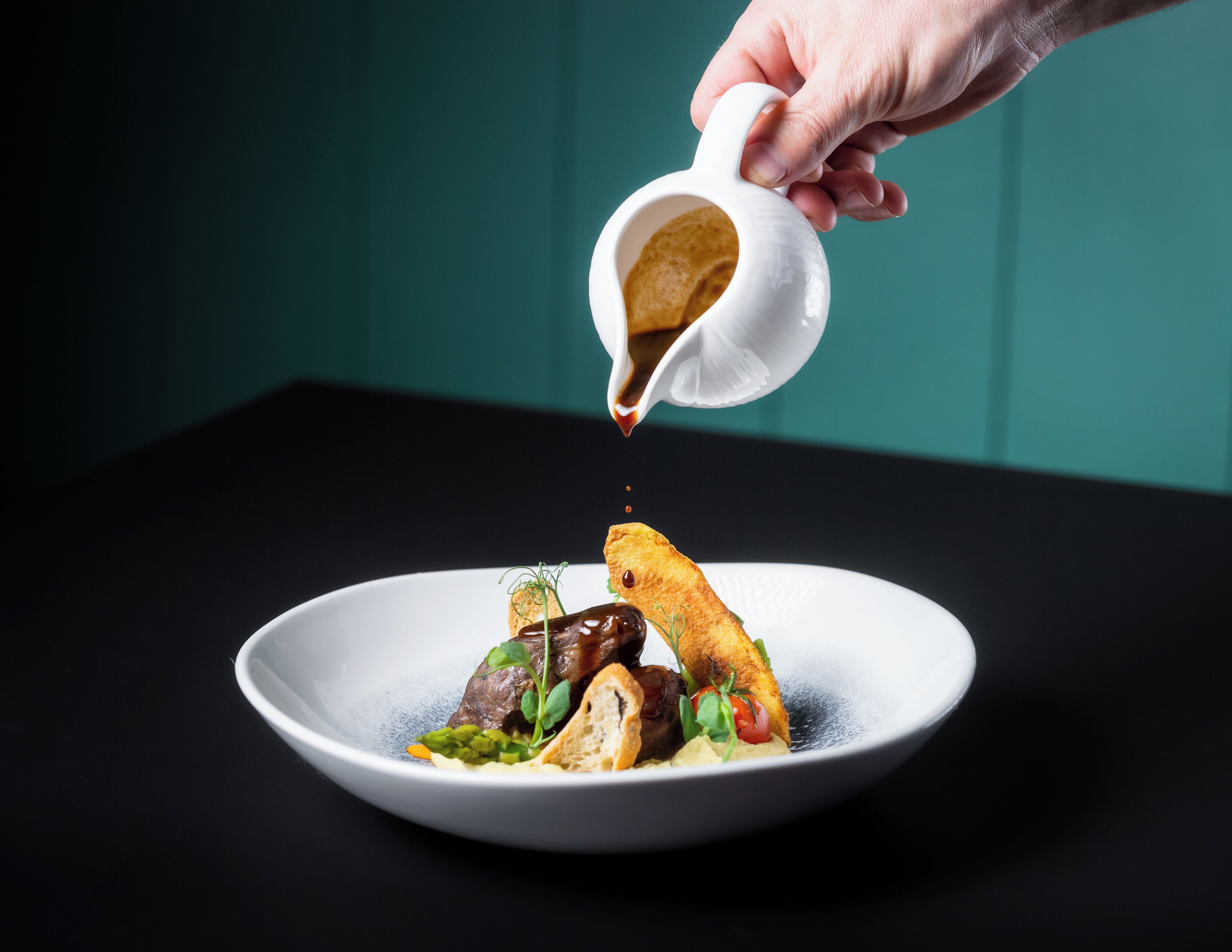 A hand pouring sauce over a dish with bread and greens.