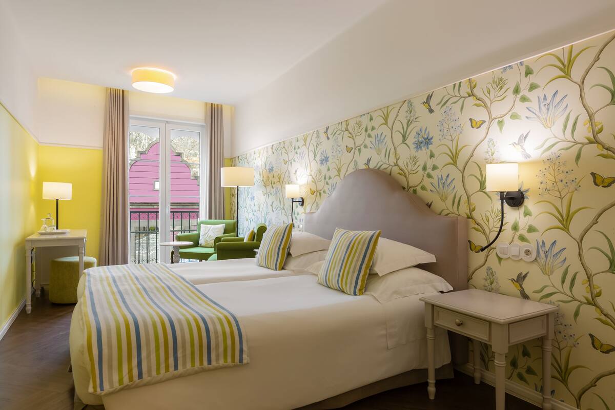 A hotel room with two beds, a small desk, a chair, and a balcony with a view.