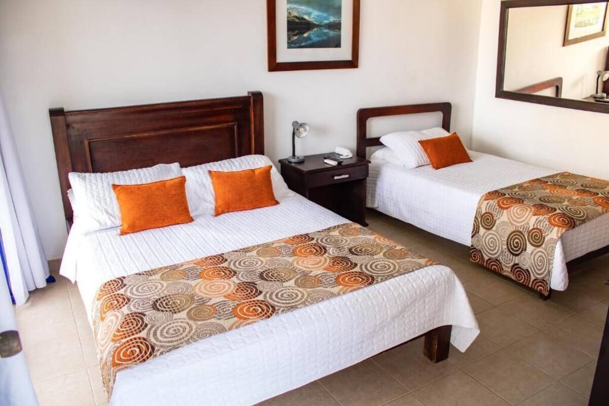 A hotel room with two single beds, each with a patterned bedspread and an orange pillow. There is a nightstand with a lamp and a phone between the beds.