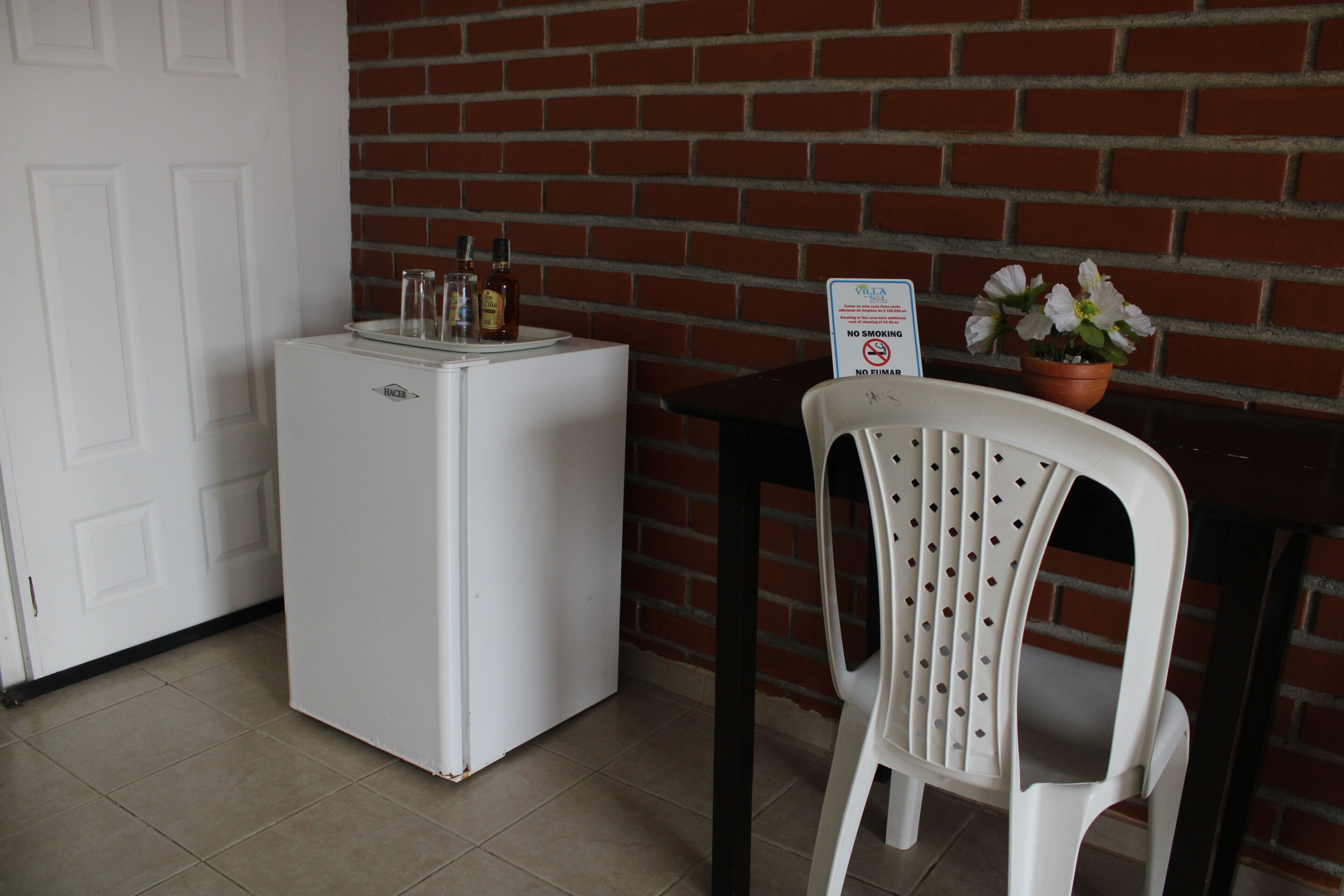 Un petit réfrigérateur blanc avec des bouteilles sur le dessus, une chaise en plastique blanche, une petite table avec un pot de plante et un panneau « interdiction de fumer ».