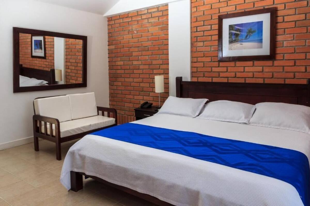 A hotel room with a brick-patterned wall, a large bed with white and blue bedding, a wooden chair, a nightstand with a lamp, and a framed picture on the wall.