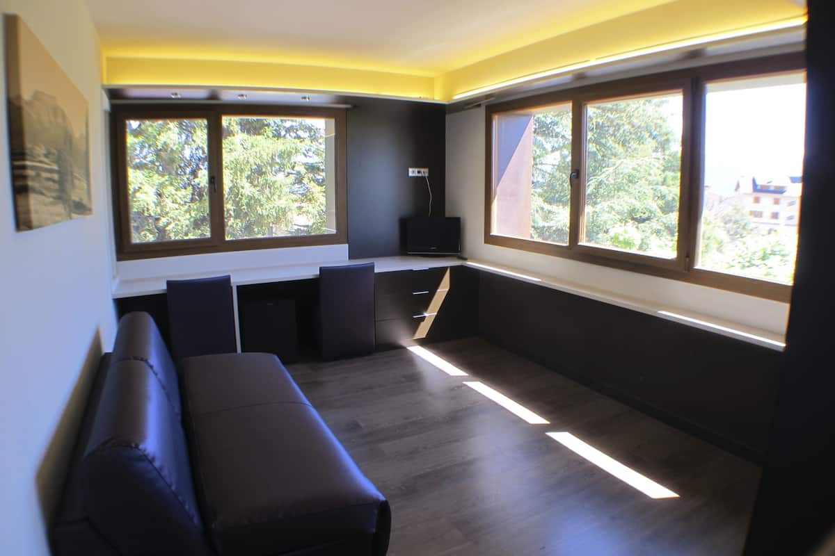A modern room with a dark-colored sofa, a flat-screen TV, and large windows offering a view of trees and buildings.