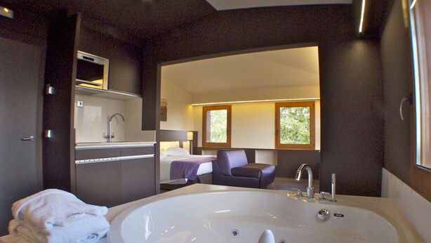 A modern bathroom with a large bathtub, a sink, and a mirror. There is a chair and a window in the background. - Hotel Mirador (Lles)