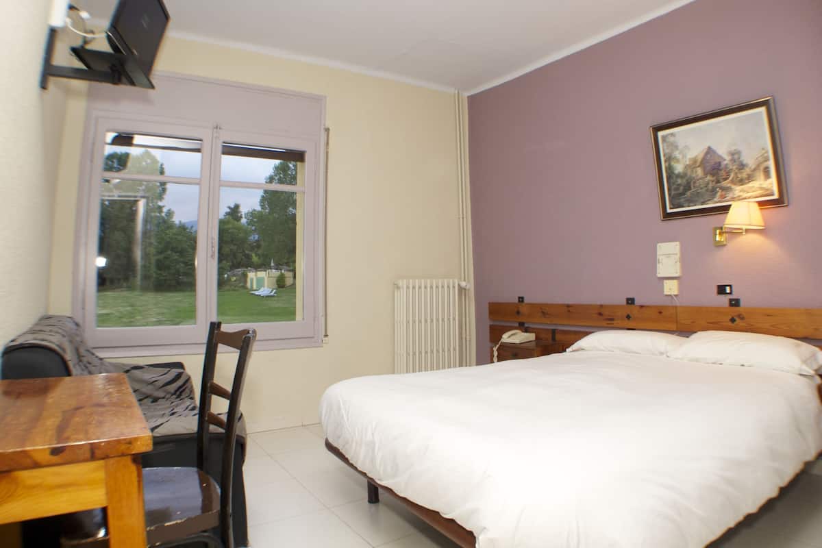 A bedroom with a bed, a desk, a chair, a window, and a painting on the wall.