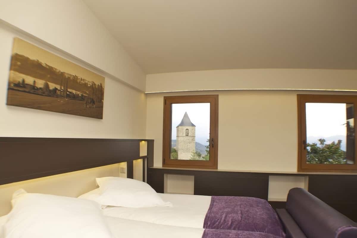 A hotel room with a bed, a view of a tower through two windows, and a painting on the wall.