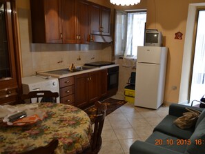Eine kleine Küche mit Holzausstattung, inklusive Herd, Spüle und Kühlschrank. Ein Esstisch mit blumigem Tischläufer und Stühlen lädt zum Verweilen ein. Im Wohnbereich ist eine Couch zu sehen.