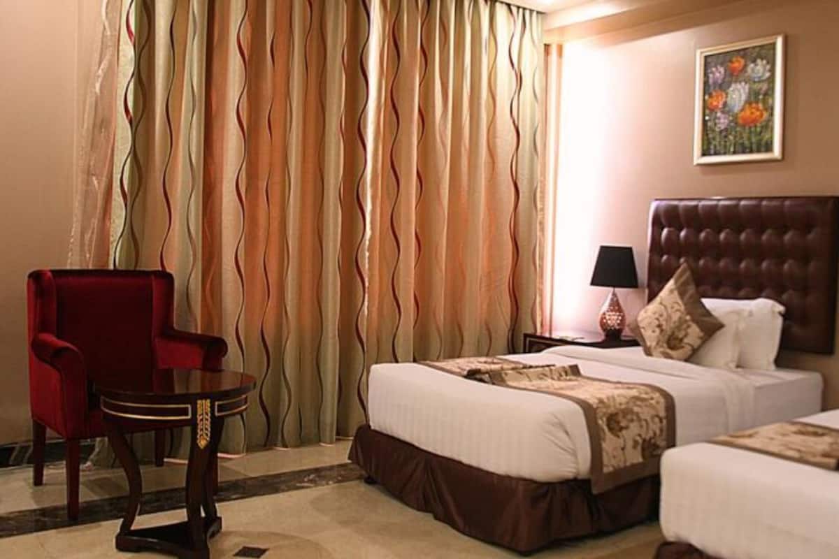 A hotel room with two beds, a red armchair, a small table, and a painting on the wall.