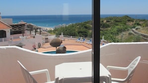 Een balkon met witte plastic stoelen en een tafel, met uitzicht op een zwembad en strand, onder helderblauwe luchten.