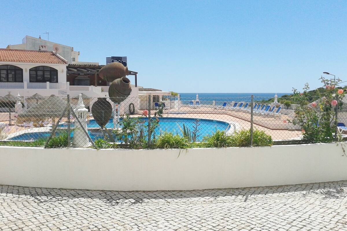 Zona de piscina con vista despejada al mar, piscina cercada y un edificio con techo de tejas.