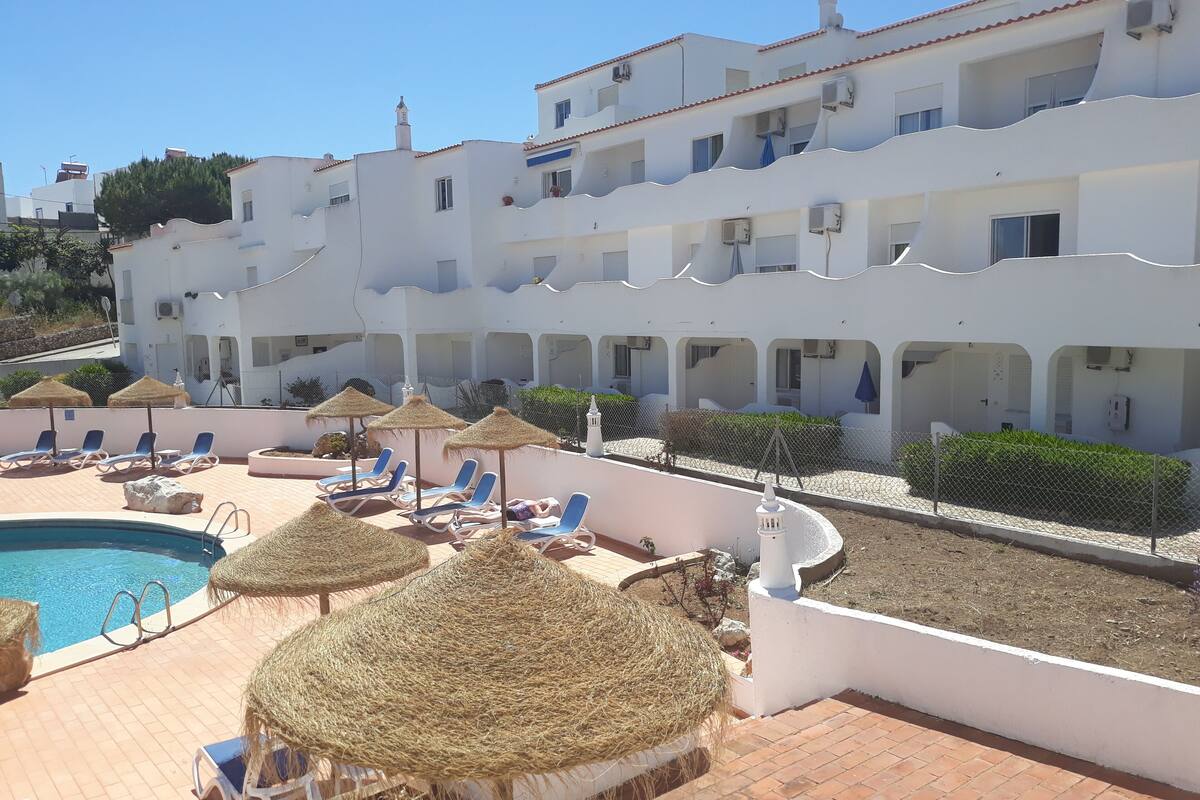 Zona de piscina con sillones de descanso y sombrillas de paja, rodeada por un edificio blanco con balcones.