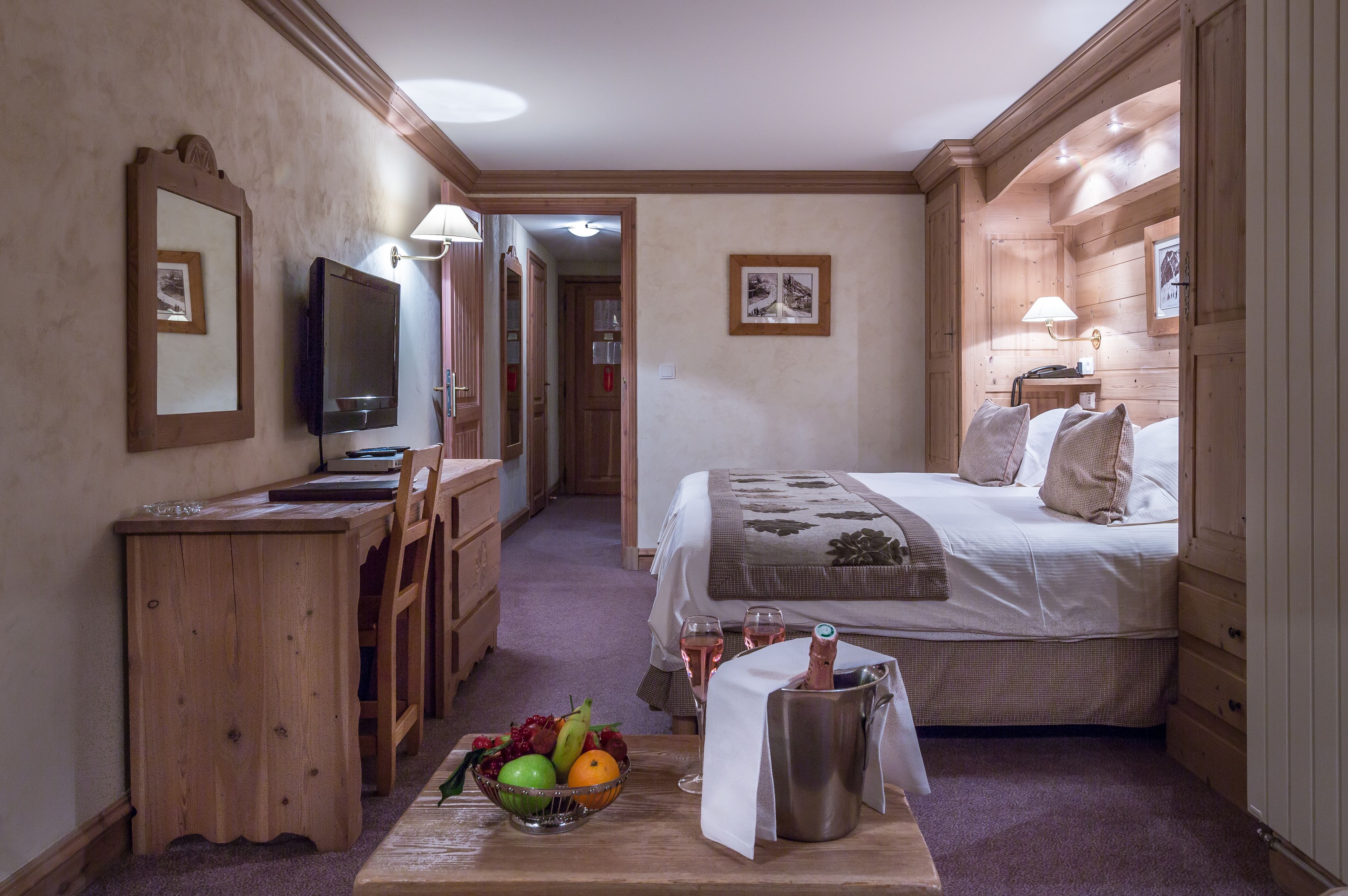 Une chambre d’hôtel avec un lit, une table de chevet, un petit bureau avec une chaise, une télévision et un panier de fruits posé sur le sol.