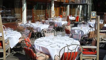 An outdoor dining area with round tables set for two, each with white tablecloths and metal chairs with red cushions.