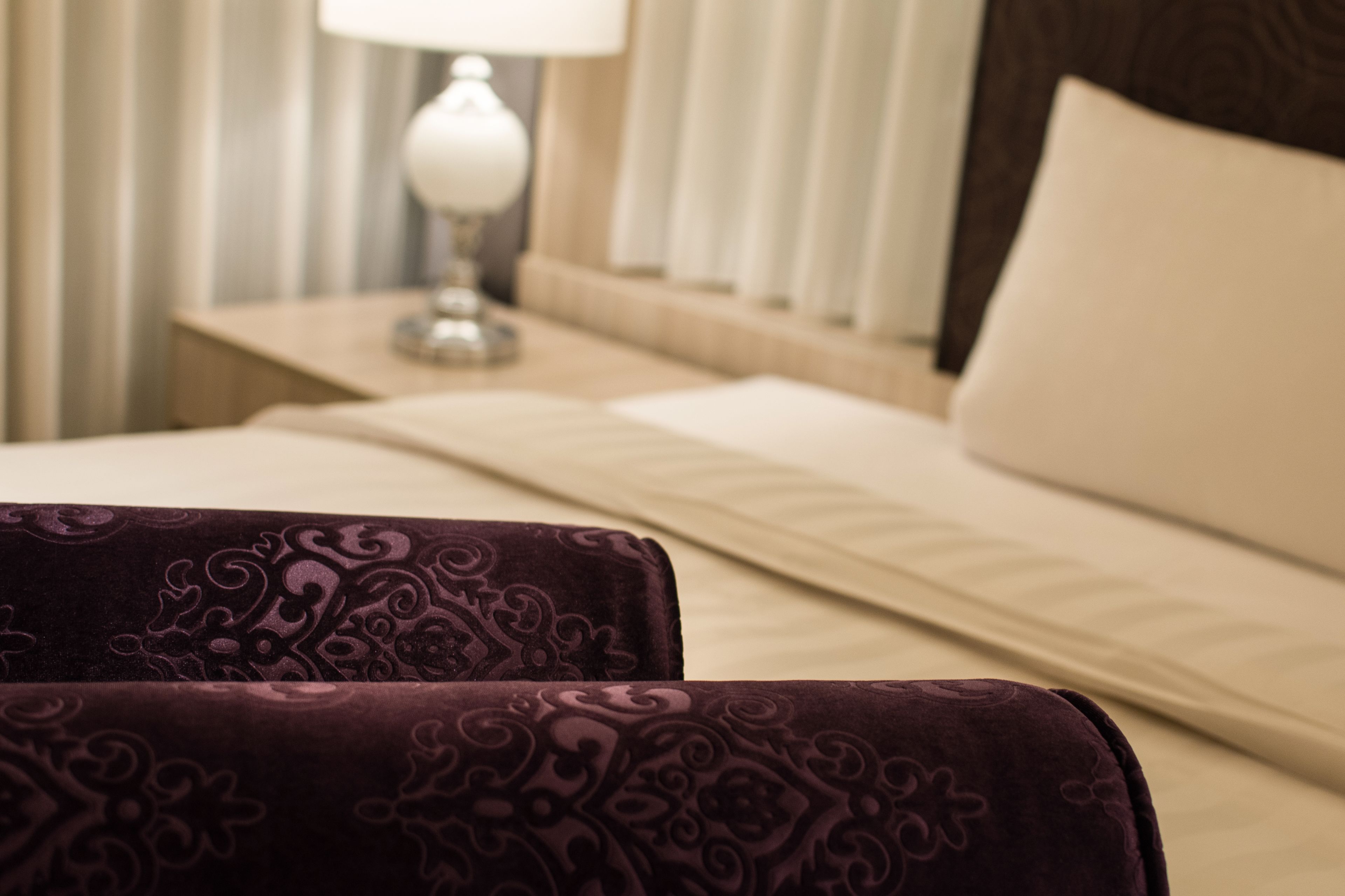 A neatly made bed with a white pillow and a headboard, a bedside table with a lamp, and a window with blinds.