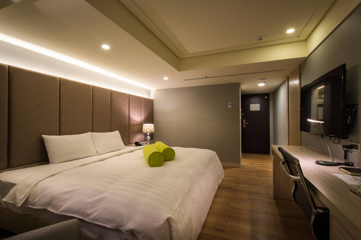A hotel room with a large bed, bedside table, lamp, and a desk with a chair.