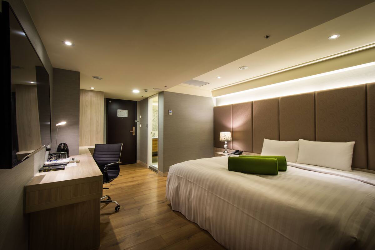 A modern hotel room with a large bed, a desk with a chair, a flat-screen TV, and a bathroom visible through an open door.