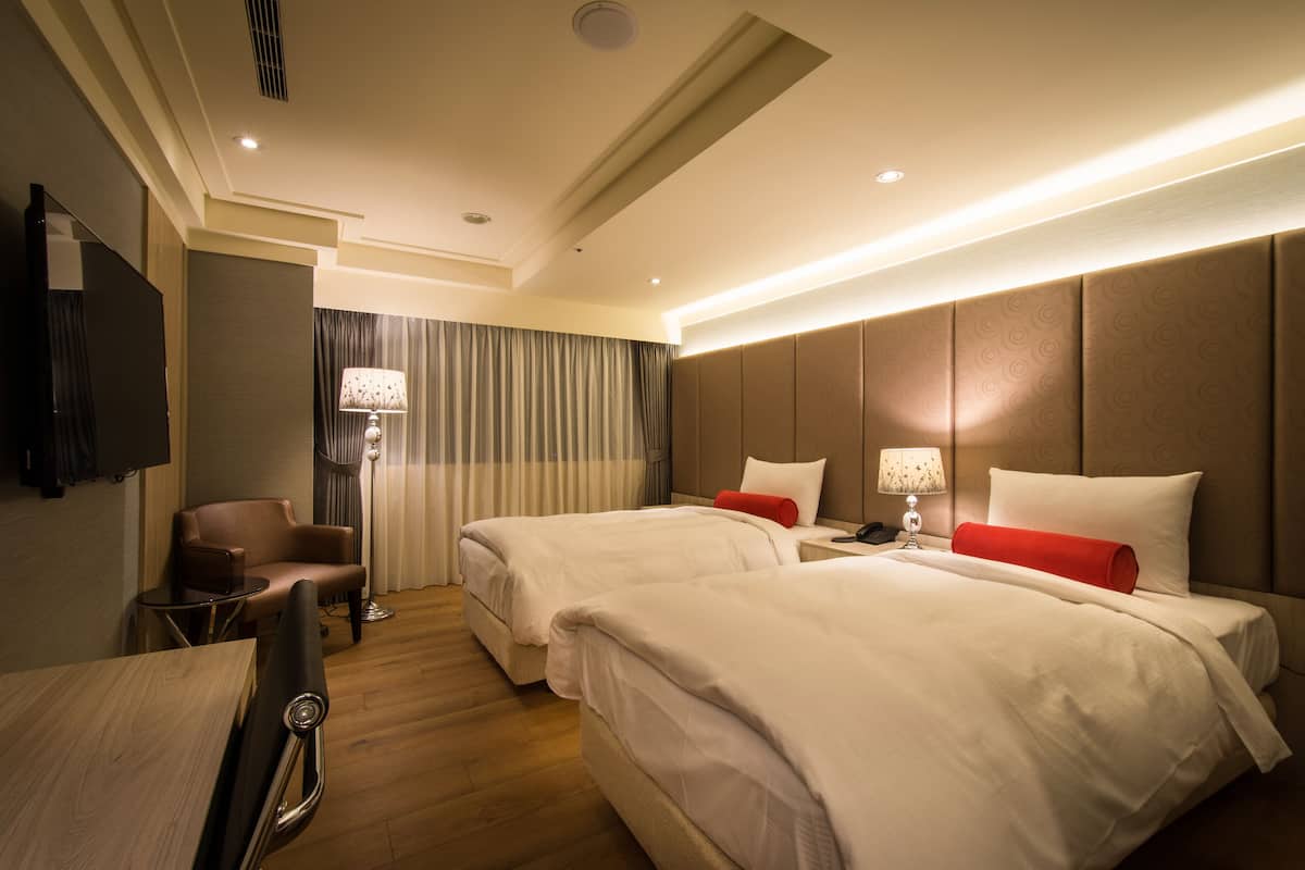 A hotel room with two beds, a desk, a chair, and a TV.