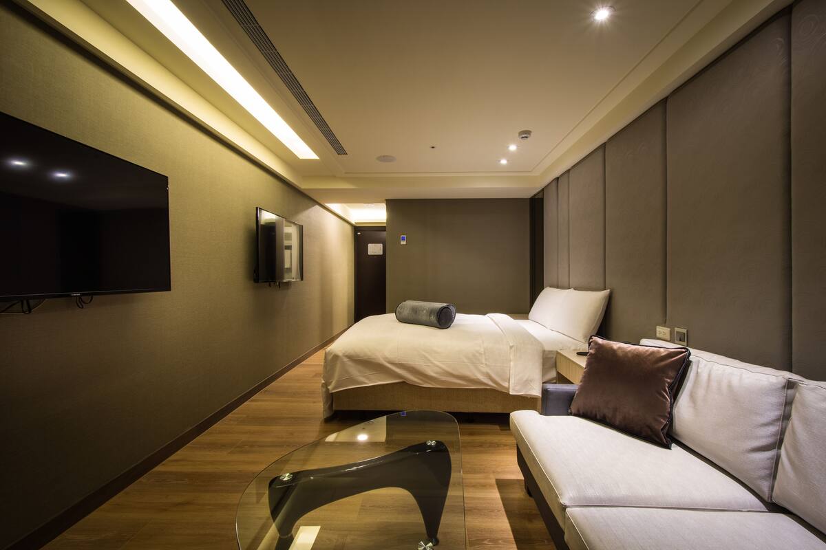 A modern hotel room with a bed, a sofa, a glass coffee table, a flat-screen TV, and a wall-mounted TV.
