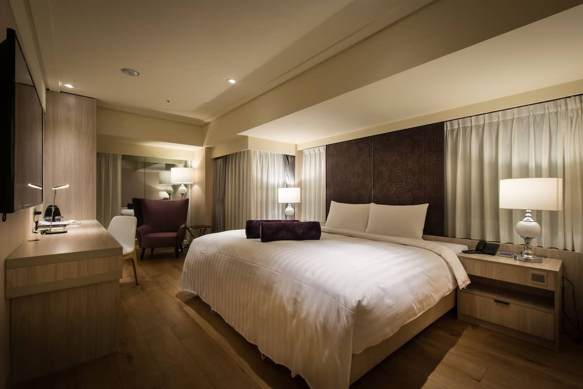 A hotel room with a large bed, a desk with a chair, a TV, and a nightstand with a lamp.