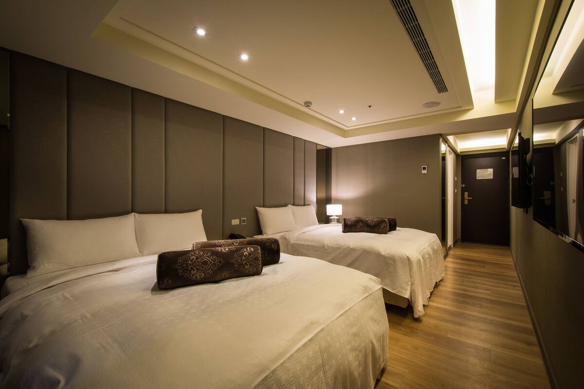 A hotel room with two beds, a wooden floor, and a hallway leading to a door.