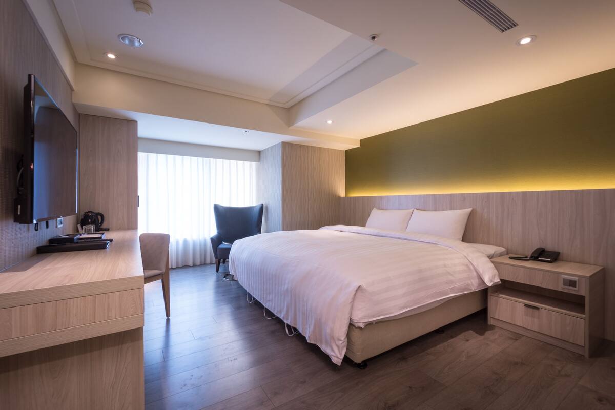 A modern hotel room with a bed, bedside table, chair, desk, and a television.
