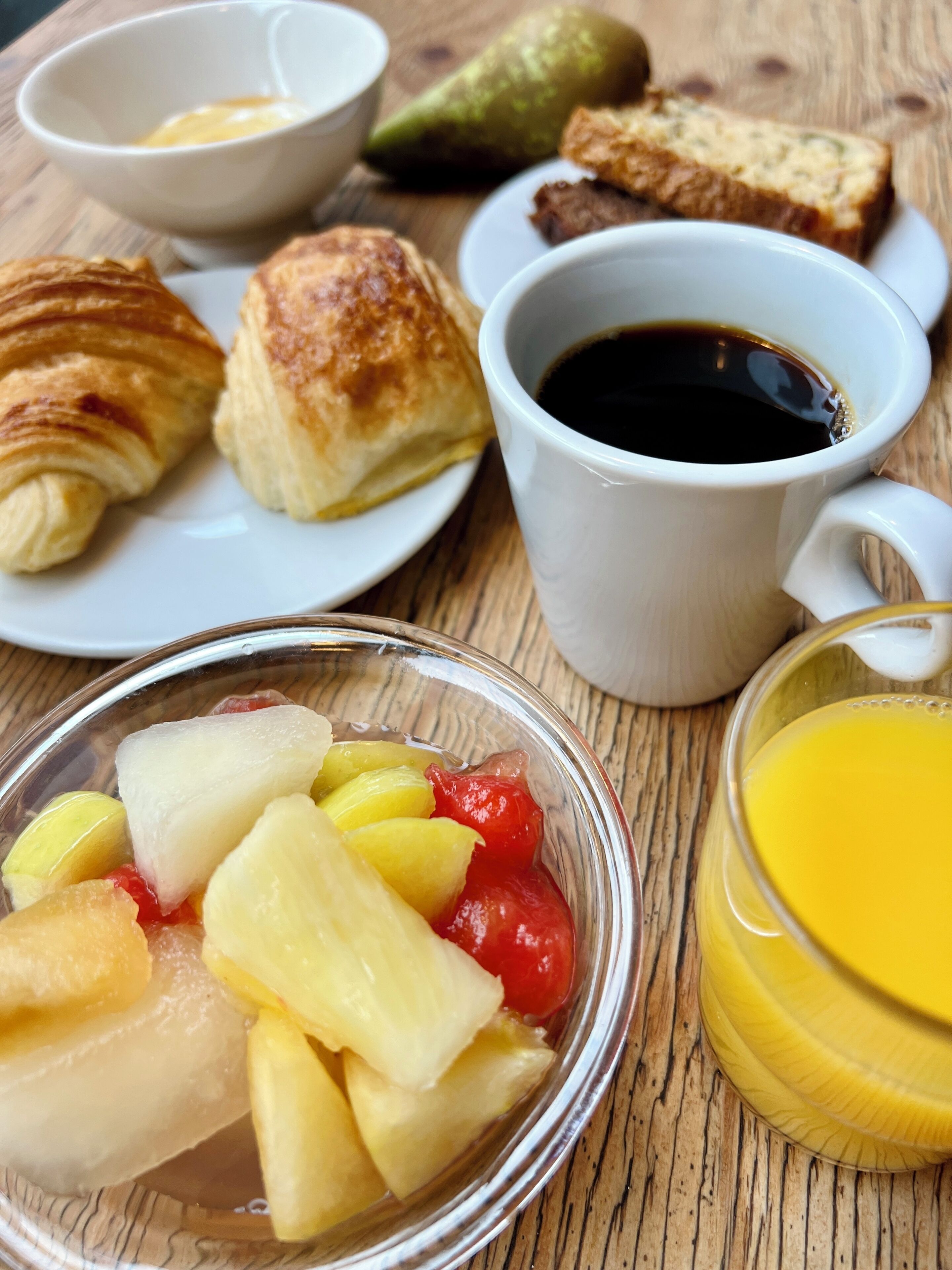 A breakfast setting with croissants, a cup of coffee, a glass of orange juice, and a bowl of fruit.