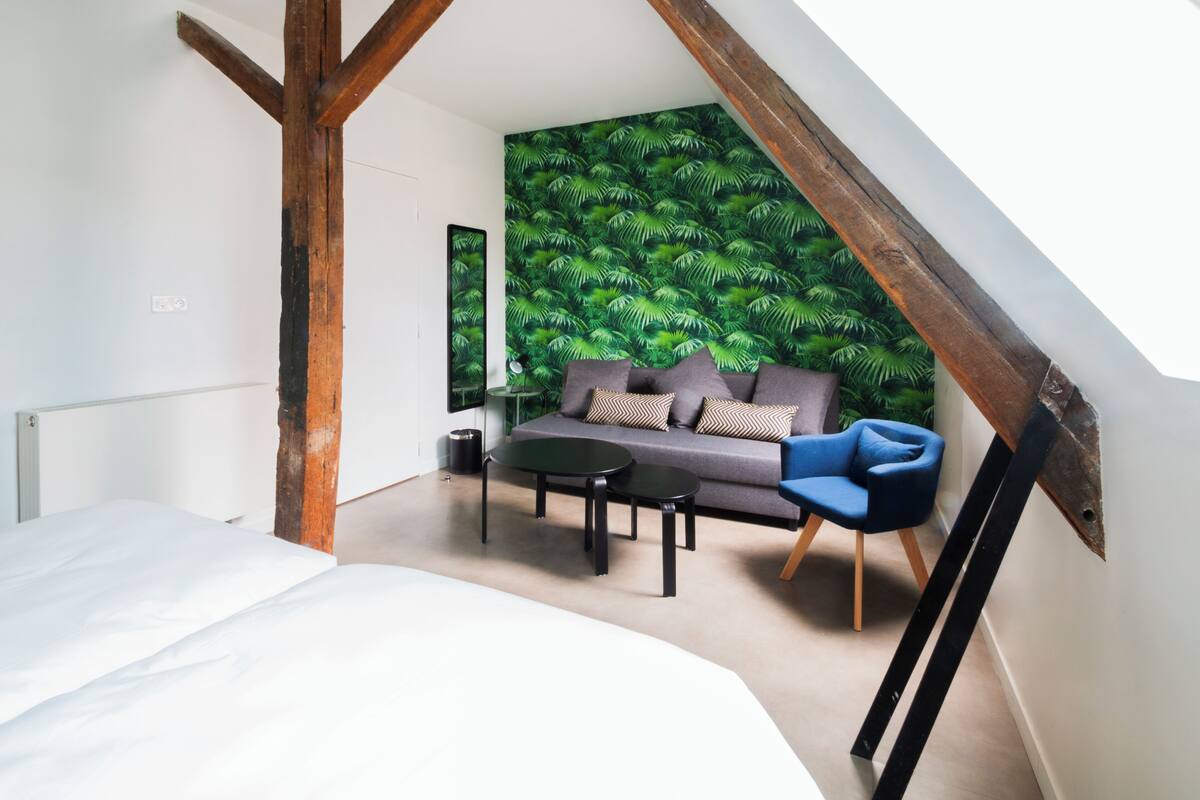 Un dormitorio con cama, sofá, dos mesitas pequeñas, un espejo y una pared con estampado de hojas verdes.