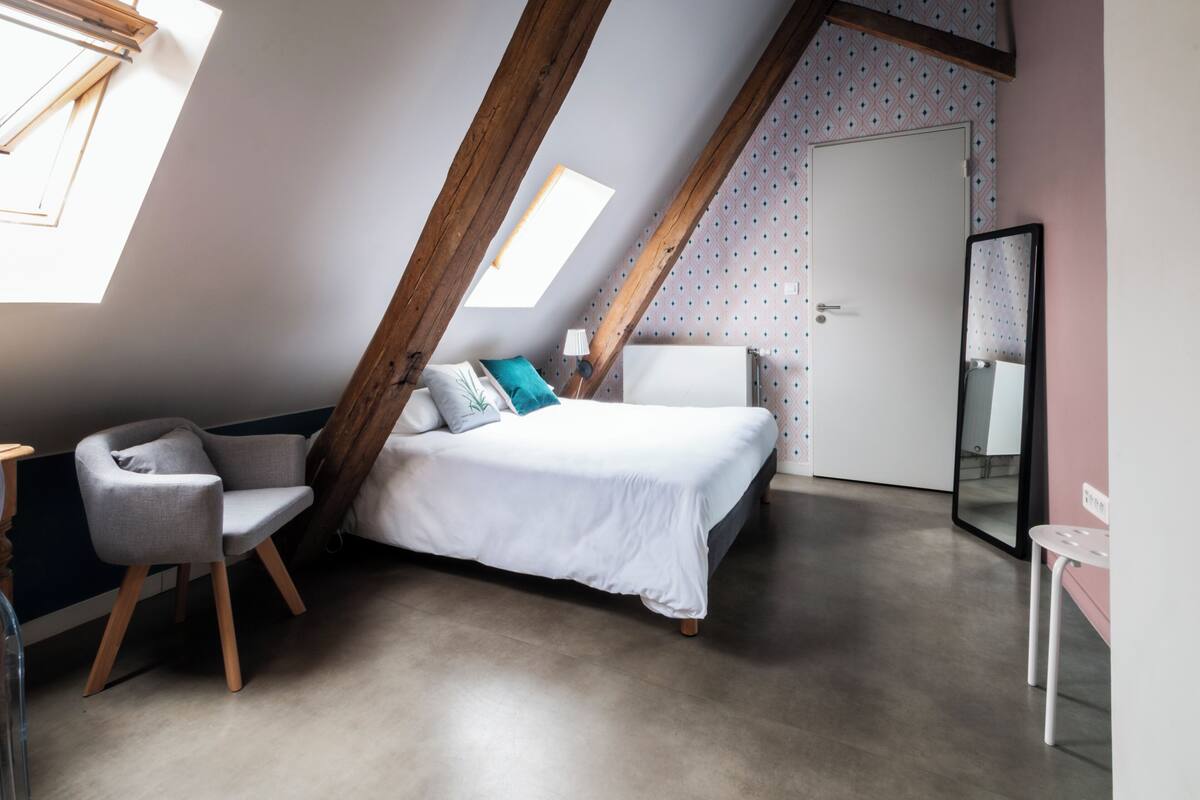 Un dormitorio con techo inclinado, una cama, una silla, un escritorio y un espejo.