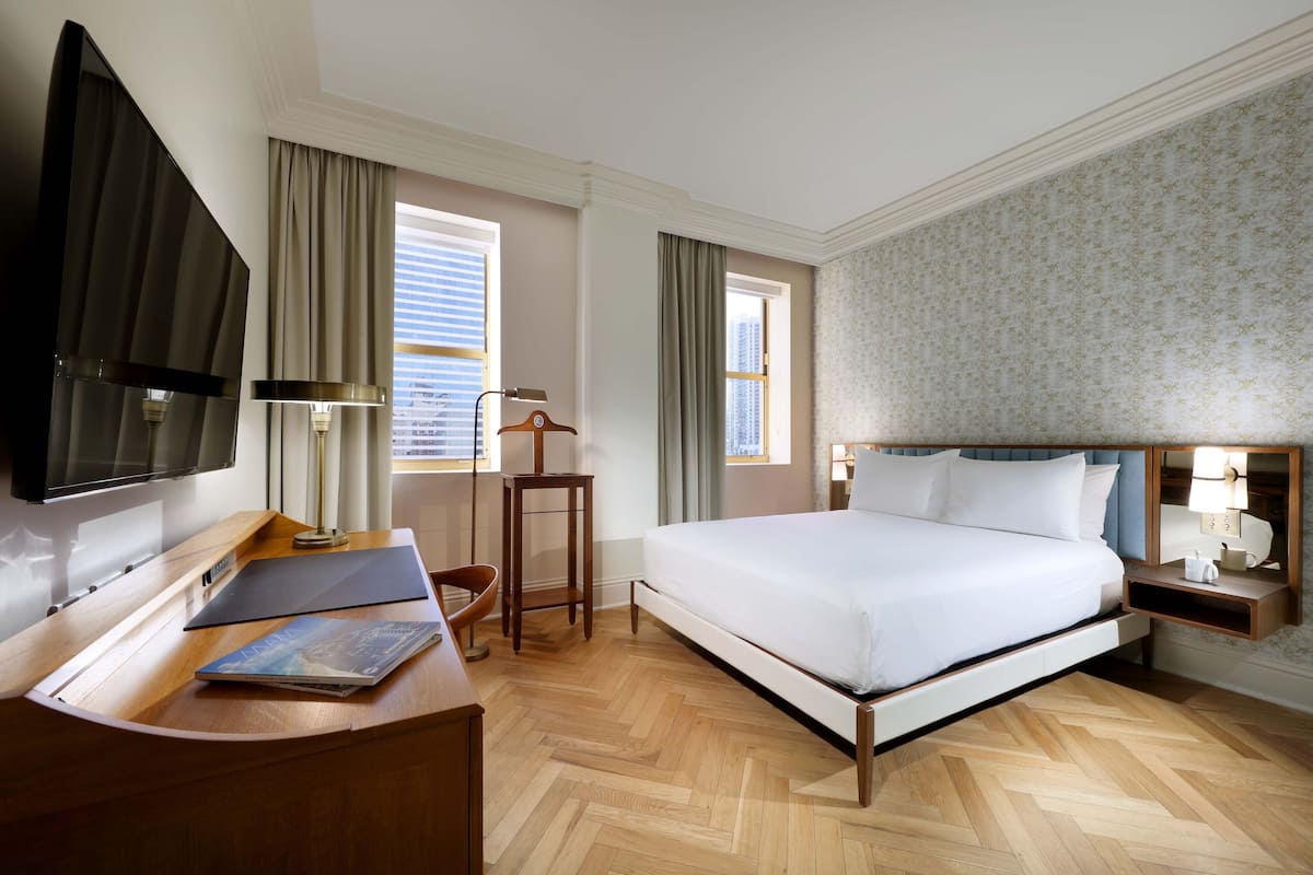 A hotel room with a bed, a desk with a chair, a TV, and a window with curtains.