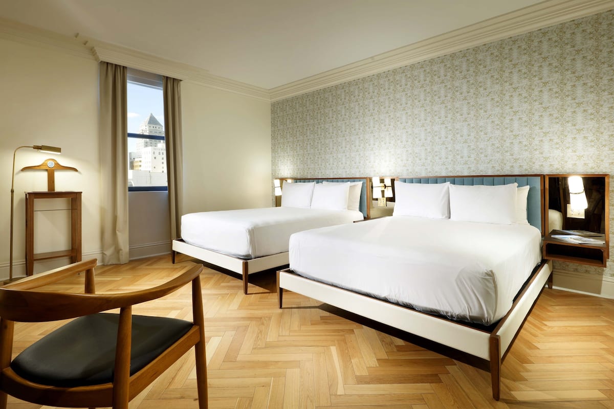A hotel room with two beds, a wooden chair, a nightstand, and a view of a building through the window.