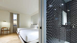 Ett hotellrum med en stor säng, en stol, ett fönster med gardiner och ett badrum med svarta kakelväggar och en duschkabin i glas.