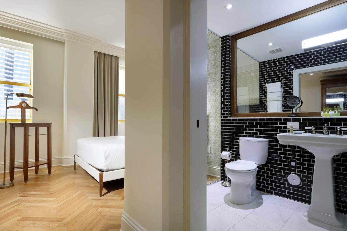 A hotel room with a bed, a wooden side table, a bathroom with a black and white tiled wall, a sink, and a mirror.