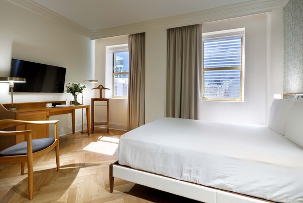 Una habitación de hotel con una cama, un escritorio, una silla, un televisor y ventanas amplias.