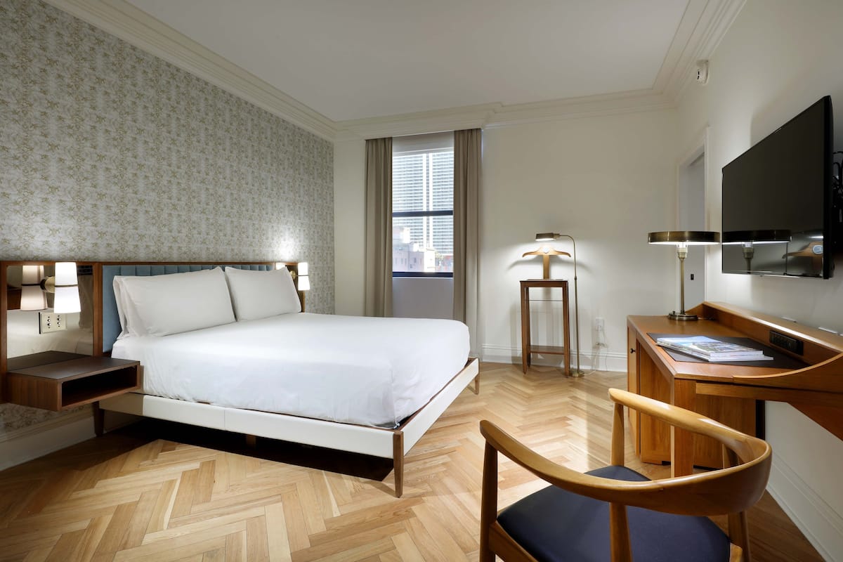 A hotel room with a bed, bedside tables, a desk, a chair, a TV, and a window with curtains.