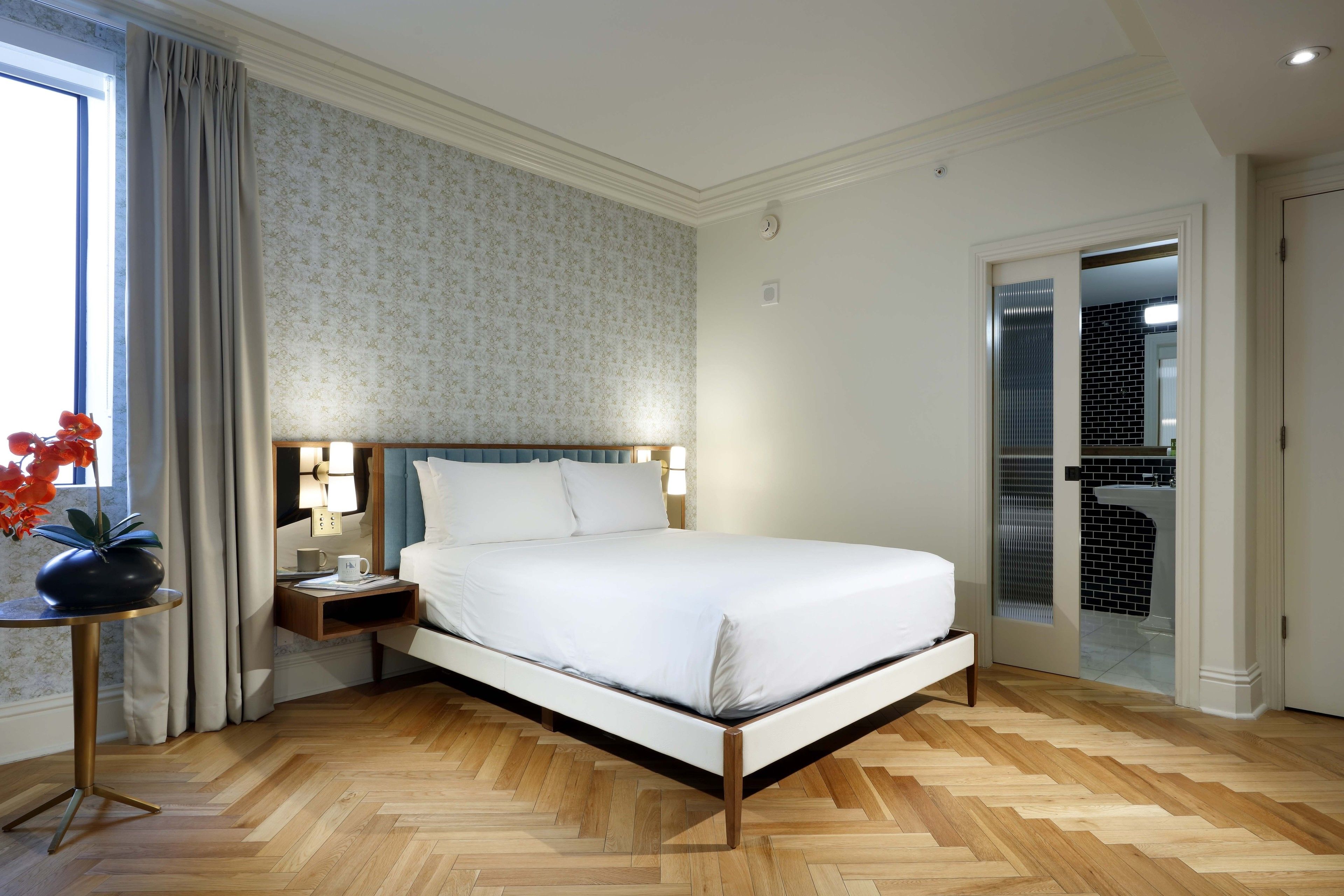 Une chambre à coucher bien rangée, avec un grand lit, une table de chevet avec une lampe, une plante en pot et une vue sur la salle de bain à travers une porte en verre.