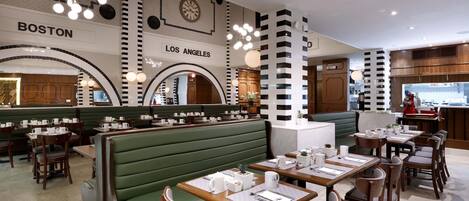 Interior de restaurante con asientos tipo cabina verdes, mesas de madera y un diseño moderno que incluye relojes y nombres de ciudades en las paredes.