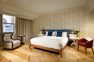 Habitación de hotel con una cama grande, una silla, una mesita de noche y vistas a un edificio desde la ventana.