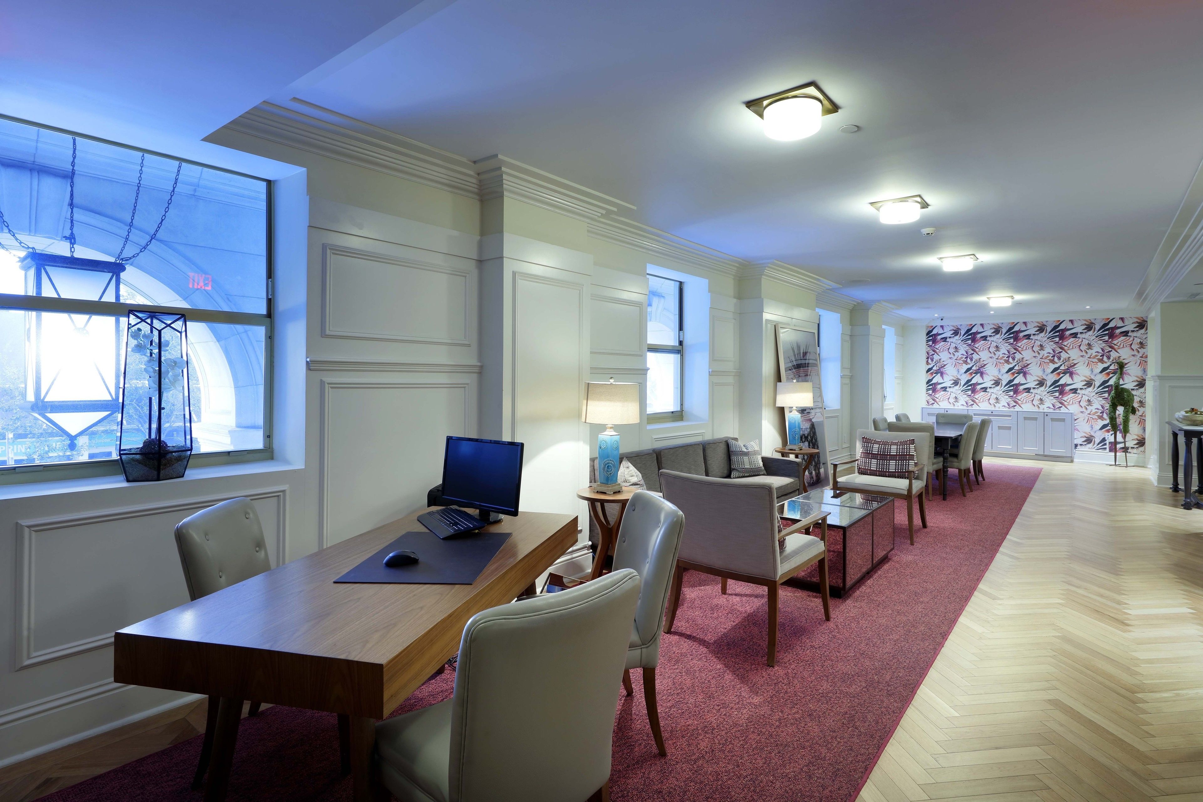 A spacious room with a large wooden desk, a computer, and a comfortable seating area with a red carpet.