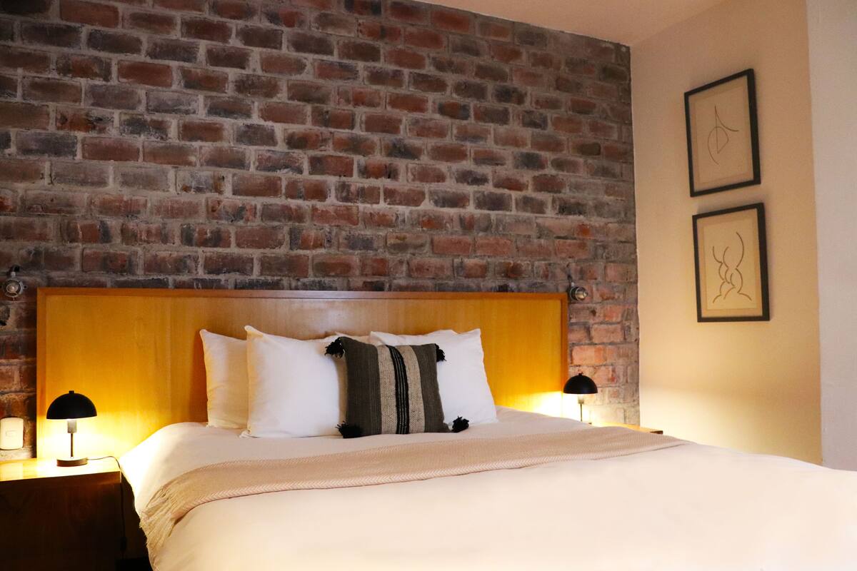 A hotel room with a brick wall, wooden headboard, white bedding, and two bedside lamps.