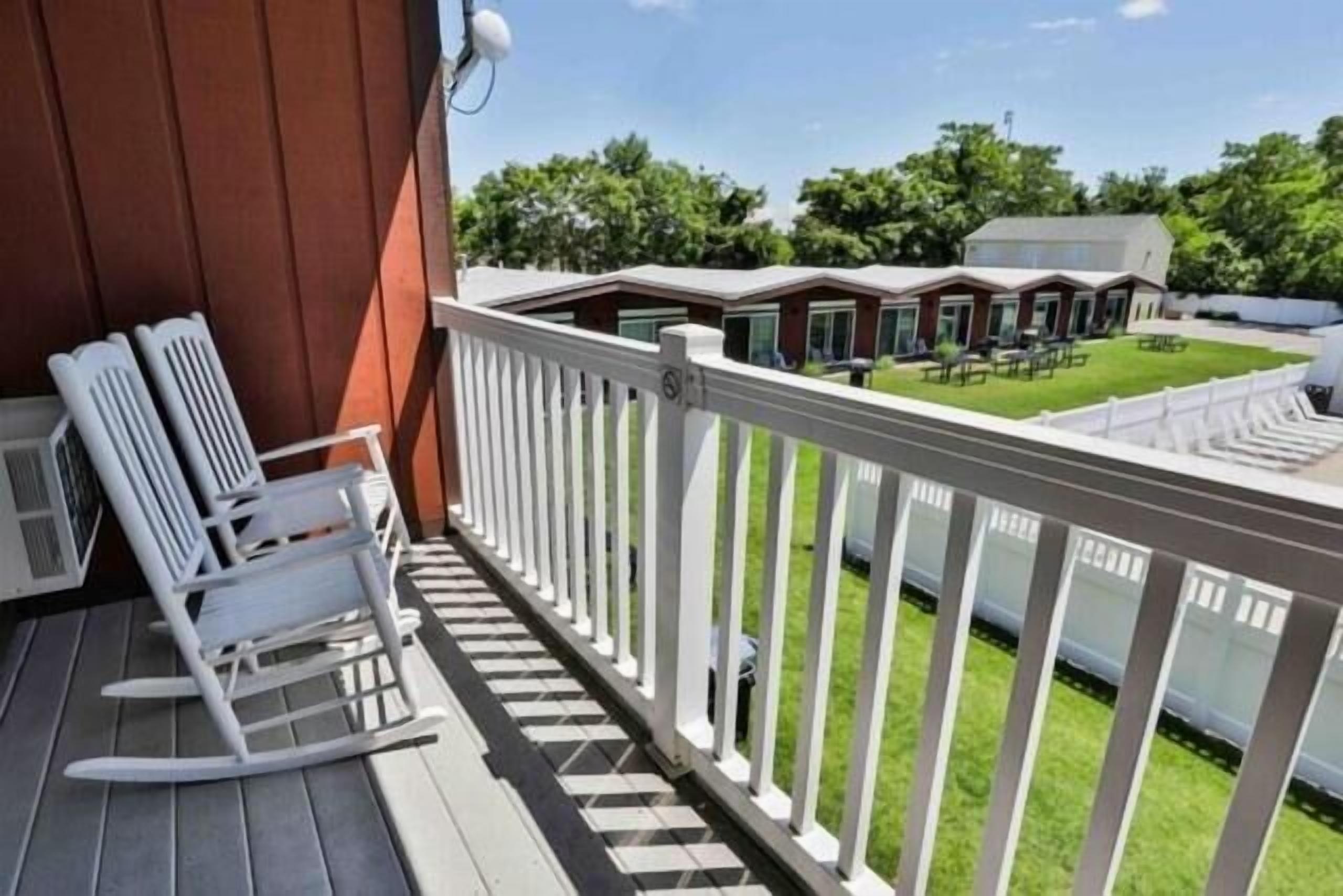 A wooden deck with white rocking chairs, a white railing, and a view of a grassy area with buildings in the background.