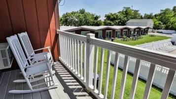 A wooden deck with white rocking chairs, a white railing, and a view of a grassy area with buildings in the background.
