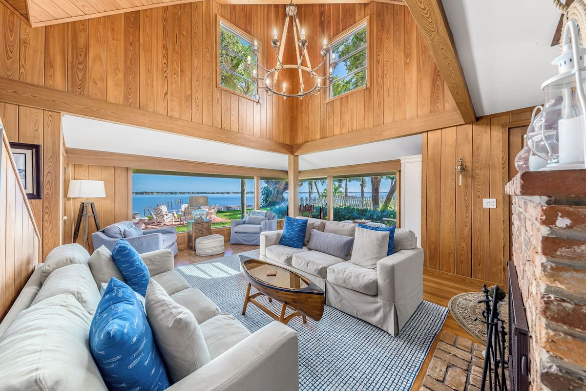 A spacious living room with a large window overlooking a body of water, wooden paneling, and a chandelier.