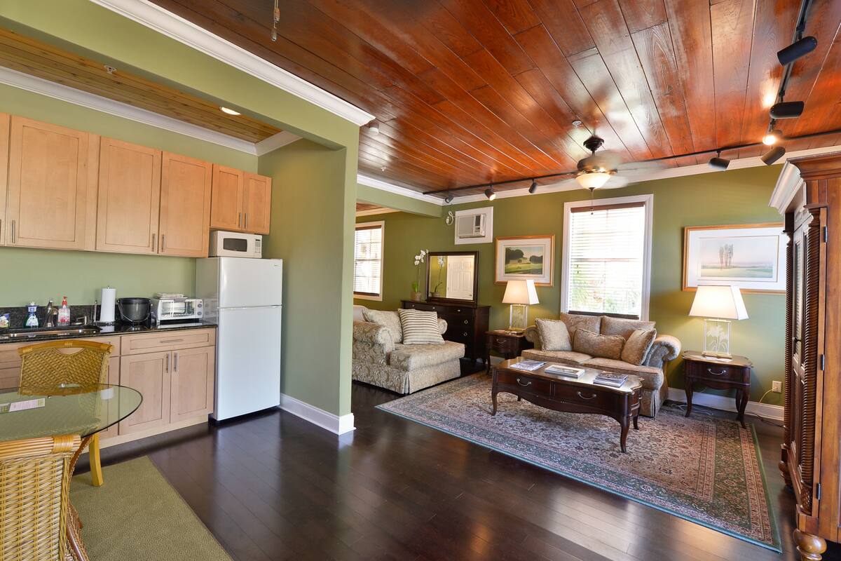 A spacious living area with a wooden ceiling, a dining table with chairs, a kitchen area with cabinets and appliances, and a cozy seating area with a rug and cushions.