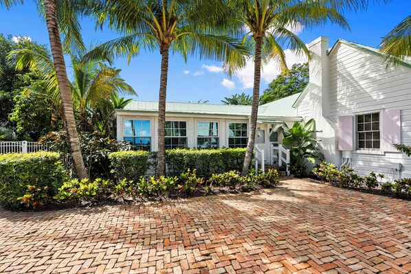 A white building with a green roof, surrounded by palm trees and a brick pathway.